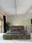 Frontale Ansicht des gemütlichen Bettes 160x200 Cloudi im olivgrünen Stoff Lumo (Chenille) in einem modernen Schlafzimmer mit Rundbogenfenster und Betonwand.