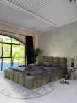 Gemütliches Schlafzimmer-Design mit dem moosgrünen Bett 180x200 Cloudi aus dem strukturierten Stoff Lumo (Chenille) neben einem großen Fenster und einer Zimmerpflanze.