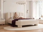 Schräge Ansicht des Designer-Polsterbettes Panola (180x200 cm) im hellen Bezug, die das Paneel-Kopfteil und die zylindrischen Beine zeigt, elegant arrangiert in einem hellen Schlafzimmer mit Stuck und einem ovalen Teppich. im RUNO_07