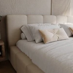 Nahaufnahme des gepolsterten Kopfteils vom Bett Ferento 140x200 cm mit Kissen, um die weiche Textur des beigen Bezugsstoffs zu verdeutlichen.