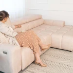 Eine entspannte Frau sitzt auf dem komfortablen beigen Ecksofa Aldo, was die gemütliche Nutzung und die praktische elektrische Relaxfunktion im Alltag demonstriert.